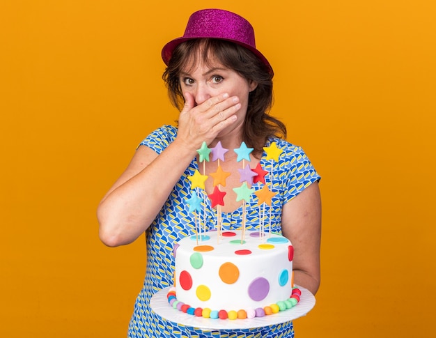 Mulher de meia-idade com um chapéu de festa segurando um bolo de aniversário em choque cobrindo a boca com a mão, comemorando a festa de aniversário em pé sobre a parede laranja