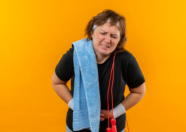 Foto grátis mulher de meia-idade, desportiva, com uma t-shirt preta com fita para a cabeça e uma toalha no ombro, parecendo indisposta, tocando a barriga, sentindo dor em pé sobre a parede laranja