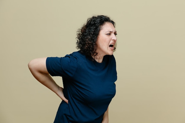 Mulher de meia-idade dolorida vestindo camiseta em vista de perfil olhando para o lado, mantendo a mão nas costas isolada em fundo verde-oliva