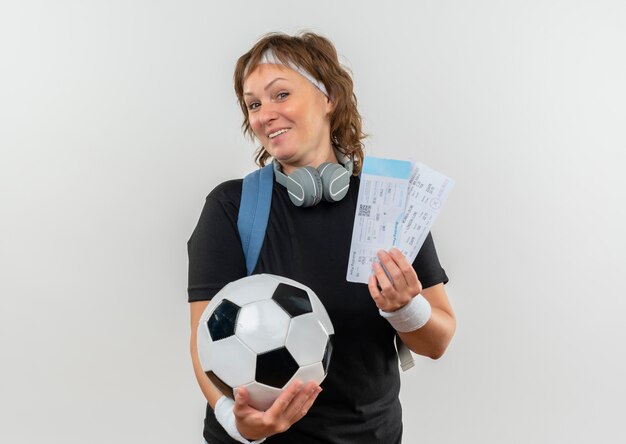 Mulher de meia-idade e esportiva em uma camiseta preta com bandana e mochila segurando passagens aéreas e uma bola de futebol sorrindo alegremente em pé sobre uma parede branca