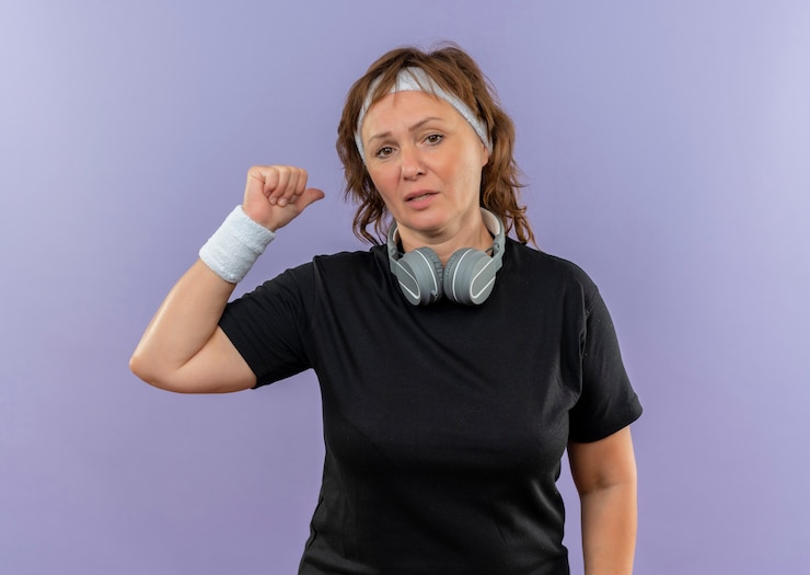 Foto grátis mulher de meia-idade, esportiva, com camiseta preta e faixa na cabeça confusa apontando com o dedo para trás em pé sobre a parede azul