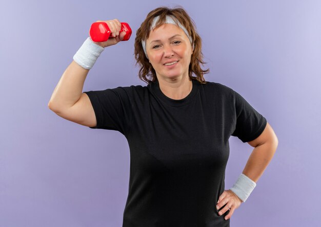 Mulher de meia idade, esportiva, com camiseta preta e faixa na cabeça, malhando com halteres, parecendo confiante em pé sobre a parede azul