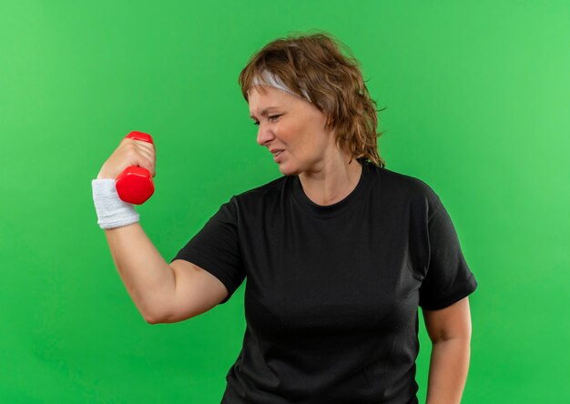 Mulher de meia-idade, esportiva, com camiseta preta e faixa na cabeça, malhando com halteres, parecendo confiante em pé sobre a parede verde