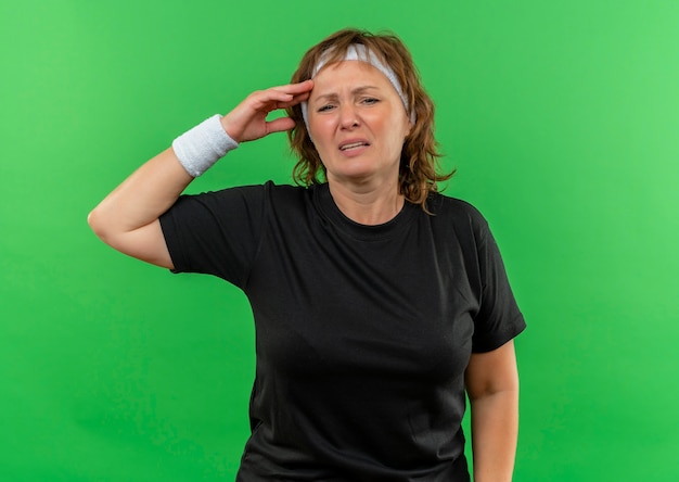 Mulher de meia-idade, esportiva, com camiseta preta e faixa na cabeça, parecendo doente e cansada em pé sobre a parede verde