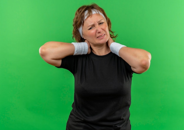 Mulher de meia-idade, esportiva, com camiseta preta e faixa na cabeça, parecendo doente, tocando o pescoço, sentindo dor em pé sobre a parede verde