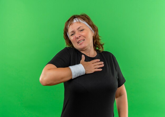 Mulher de meia-idade, esportiva, com camiseta preta e faixa na cabeça, segurando a mão no peito, parecendo indisposta em pé sobre a parede verde