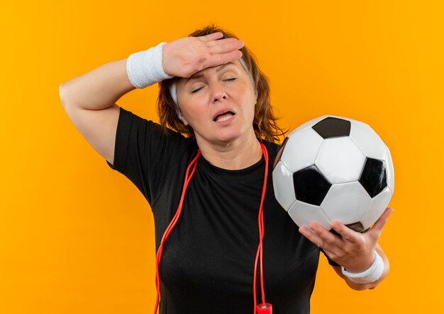 Mulher de meia-idade, esportiva, com camiseta preta e faixa na cabeça, segurando uma bola de futebol, parecendo cansada e sobrecarregada de pé sobre a parede laranja