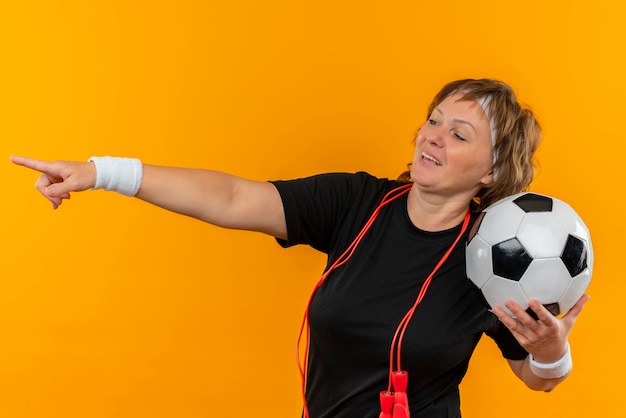 Mulher de meia-idade, esportiva, com camiseta preta e fita na cabeça, segurando uma bola de futebol apontando com o dedo para o lado e sorrindo em pé sobre a parede laranja