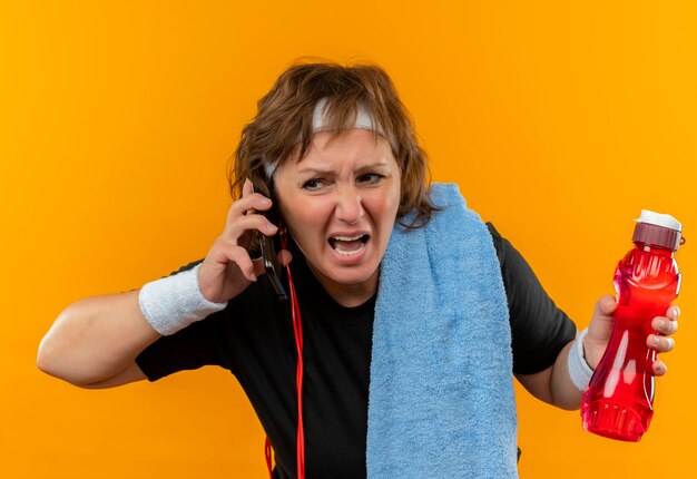 Mulher de meia-idade, esportiva, com camiseta preta, faixa na cabeça e toalha no ombro, falando no celular, parecendo decepcionada, gritando com expressão agressiva em pé sobre a parede laranja