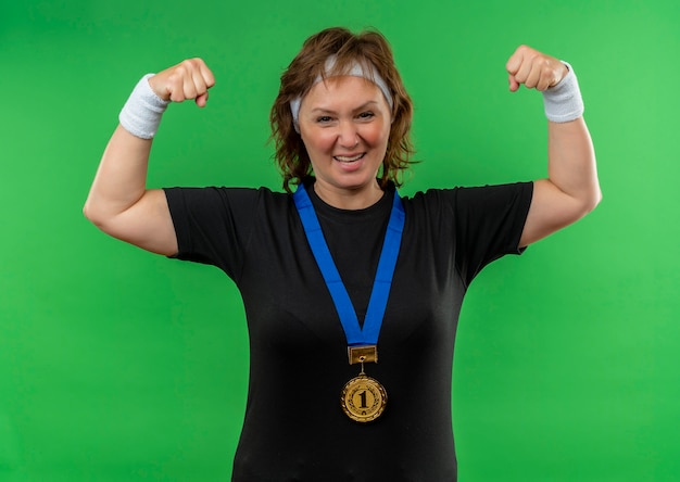 Foto grátis mulher de meia-idade, esportiva, com camiseta preta, faixa para a cabeça e medalha de ouro em volta do pescoço, cerrando os punhos, feliz e animada em pé sobre a parede verde