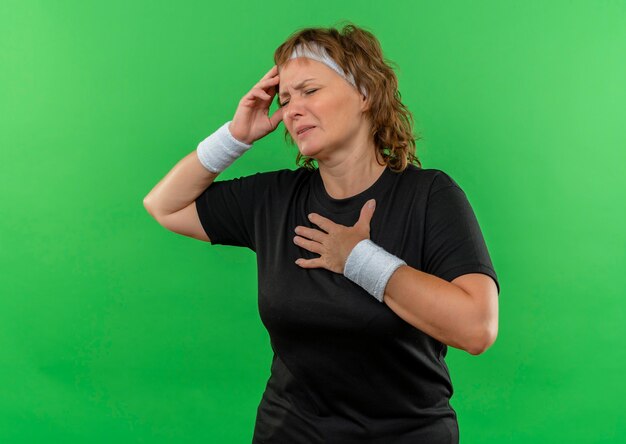 Mulher de meia-idade, esportiva, com uma camiseta preta e uma faixa na cabeça, parecendo doente, tocando sua cabeça, sentindo-se enjoada e cansada em pé sobre uma parede verde