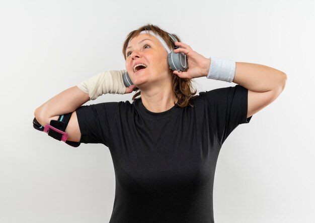 Mulher de meia-idade, esportiva, vestindo uma camiseta preta com bandana e fones de ouvido, curtindo sua música favorita, feliz e positiva, sorrindo em pé sobre uma parede branca