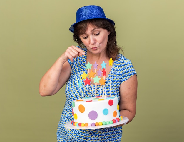 Mulher de meia-idade feliz com um chapéu de festa segurando um bolo de aniversário olhando intrigada para ele