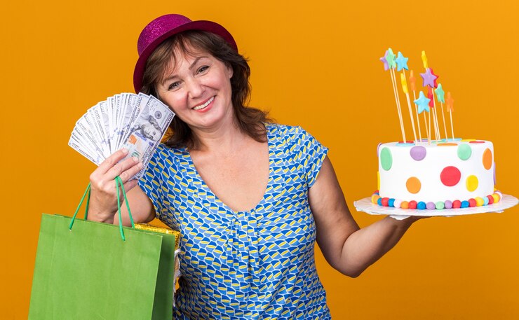Foto grátis mulher de meia-idade feliz com um chapéu de festa segurando um saco de papel com presentes segurando um bolo de aniversário e dinheiro sorrindo amplamente