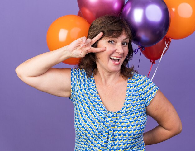 Mulher de meia-idade feliz com um monte de balões coloridos mostrando o sinal V sorrindo alegremente