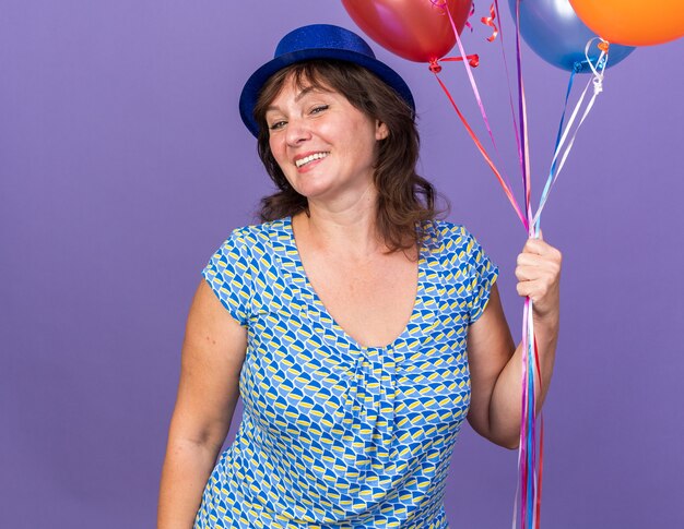 Mulher de meia-idade feliz e alegre com um chapéu de festa segurando um monte de balões coloridos sorrindo amplamente, comemorando a festa de aniversário em pé sobre a parede roxa