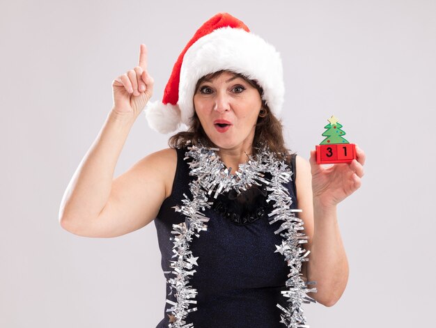 Mulher de meia-idade impressionada com chapéu de Papai Noel e guirlanda de ouropel no pescoço segurando um brinquedo de árvore de Natal com data olhando para a câmera apontando para cima, isolado no fundo branco