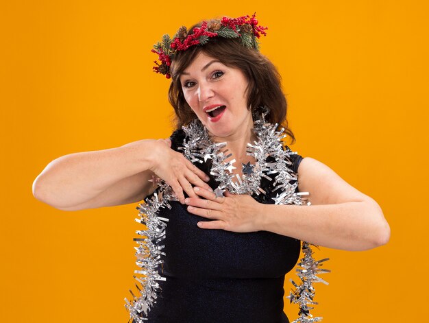 Mulher de meia-idade impressionada com coroa de flores de Natal e guirlanda de ouropel no pescoço, olhando para a câmera e mantendo as mãos no peito isoladas em fundo laranja