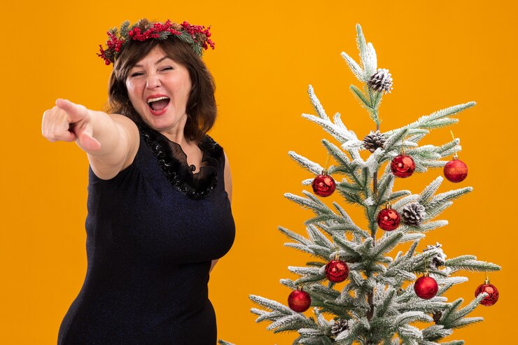 Foto grátis mulher de meia-idade impressionada com uma coroa de natal na cabeça e uma guirlanda de ouropel em volta do pescoço, perto de uma árvore de natal decorada