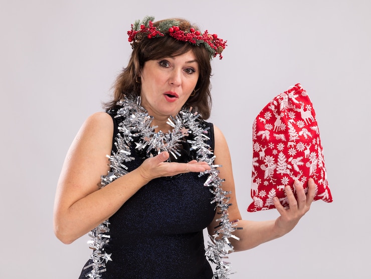 Foto grátis mulher de meia-idade impressionada usando coroa de flores de natal e guirlanda de ouropel em volta do pescoço segurando e apontando para um saco de presente de natal, olhando para a câmera isolada no fundo branco