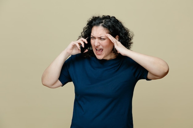 Mulher de meia idade irritada vestindo camiseta mantendo a mão na cabeça olhando para o lado gritando enquanto falava no telefone isolado em fundo verde oliva