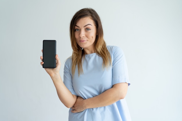 Mulher de meia idade positiva que mostra a tela do smartphone e que olha a câmera.
