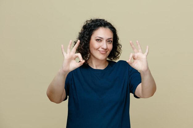 Mulher de meia idade satisfeita vestindo camiseta olhando para a câmera mostrando sinal de ok com as duas mãos isoladas em fundo verde-oliva