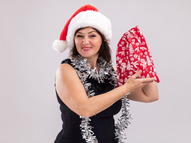 Mulher de meia-idade sorridente com chapéu de Papai Noel e guirlanda de ouropel no pescoço segurando um saco de presente de Natal, olhando para a câmera, isolada no fundo branco com espaço de cópia