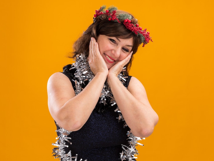 Foto grátis mulher de meia-idade sorridente com coroa de natal na cabeça e guirlanda de ouropel no pescoço, olhando para a câmera, mantendo as mãos no rosto isoladas em um fundo laranja com espaço de cópia