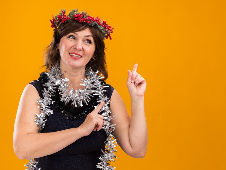 Foto grátis mulher de meia-idade sorridente usando coroa de natal na cabeça e guirlanda de ouropel no pescoço, olhando para o lado apontando para cima isolado em um fundo laranja com espaço de cópia