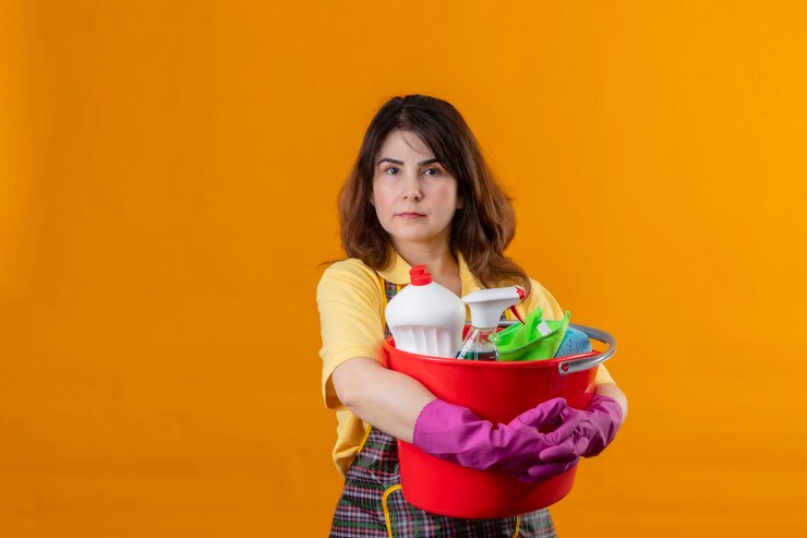 Foto grátis mulher de meia-idade usando avental e luvas de borracha segurando um balde com ferramentas de limpeza e uma expressão de confiança séria no rosto em pé sobre a parede laranja