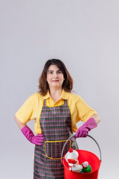 Mulher de meia-idade usando avental e luvas de borracha, segurando um balde com ferramentas de limpeza, olhando para a câmera com uma expressão séria e confiante no rosto, sorrindo amigável, pronta para limpar sobre fundo branco