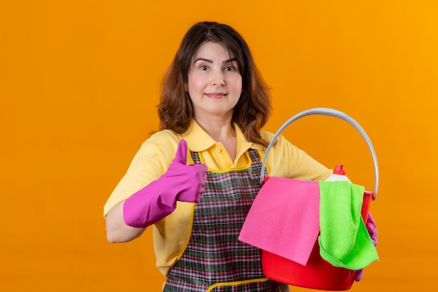 Mulher de meia-idade usando avental e luvas de borracha, segurando um balde com ferramentas de limpeza, olhando para a câmera, sorrindo alegremente positivo e feliz levantando o punho, regozijando-se com seu sucesso sobre fundo laranja