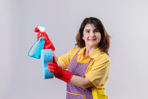 Mulher de meia-idade usando avental e luvas de borracha segurando um spray de limpeza e uma esponja