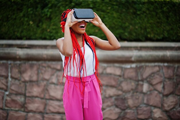 Mulher de negócios afro-americana na moda em calças cor de rosa e dreads vermelhos com óculos de realidade virtual ao ar livre