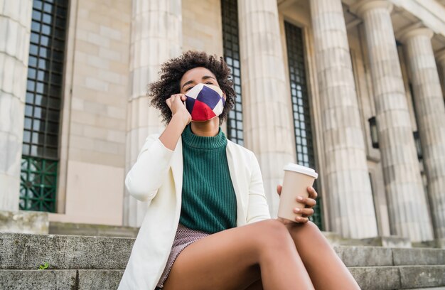 Mulher de negócios afro usando máscara protetora e falando ao telefone enquanto está sentado na escada ao ar livre na rua. Negócios e conceito urbano.