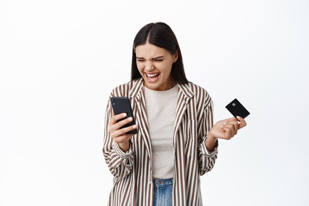 Mulher de negócios animada sorrindo e olhando para o smartphone, ganhando dinheiro online, segurando um cartão de crédito de plástico, verificando a conta bancária no aplicativo