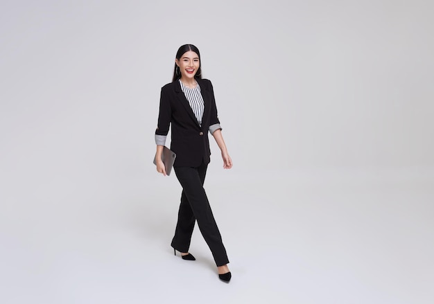 Mulher de negócios asiática feliz sorrindo em traje formal segurando tablet e caminhando sobre fundo cinza