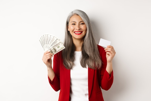 Mulher de negócios asiática sênior bem-sucedida mostrando dinheiro em dólares e cartão de plástico, sorrindo feliz para a câmera, vestindo blazer vermelho e maquiagem