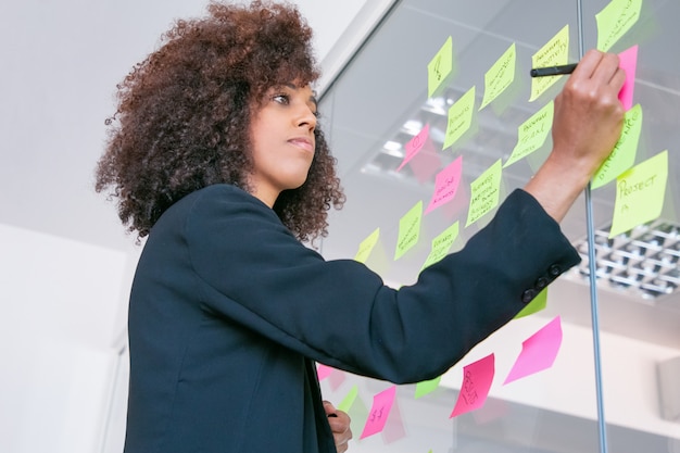 Foto grátis mulher de negócios bonita bem sucedida escrevendo na etiqueta com marcador. gerente feminina encaracolada confiante pensativa, compartilhando a ideia para o projeto e fazendo anotações. brainstorming, negócios e conceito de treinamento