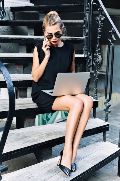 Mulher de negócios bonita em vestido preto curto, sentado na escada ao ar livre. Ela está trabalhando com um laptop, falando ao telefone.