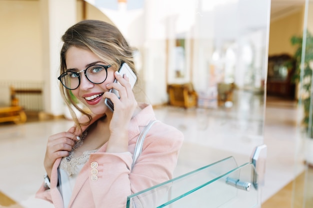 Mulher de negócios bonita, jovem sorrindo e falando por telefone, olhando, em pé no corredor. Vestindo uma elegante jaqueta rosa, óculos. Imagem através da porta de vidro.