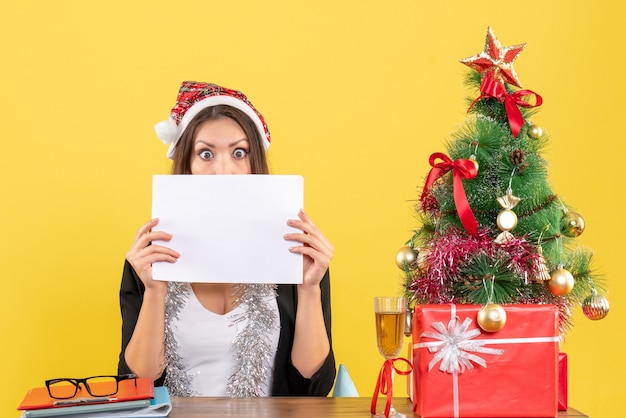 Mulher de negócios chocada, de terno com chapéu de papai noel e decorações de ano novo, trabalhando sozinha e sentada em uma mesa com uma árvore de natal no escritório