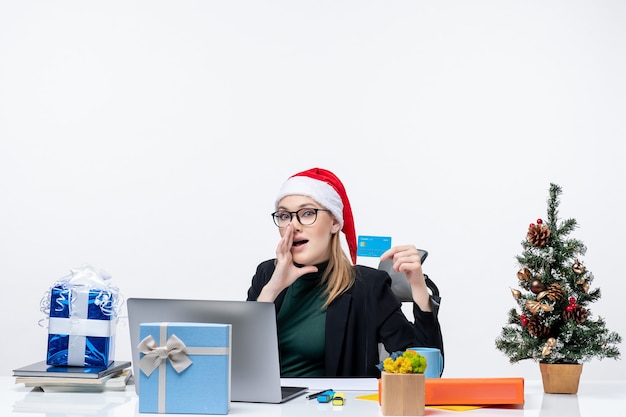 Mulher de negócios com chapéu de papai noel e usando óculos, sentada à mesa segurando um cartão de banco e ligando para alguém no escritório