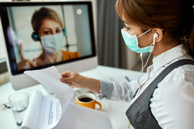 Mulher de negócios com máscara facial analisando a papelada enquanto faz videoconferência no pc de mesa no escritório