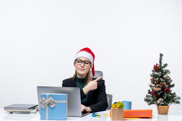 Mulher de negócios com um chapéu de papai noel sentada à mesa com uma árvore de natal e um presente