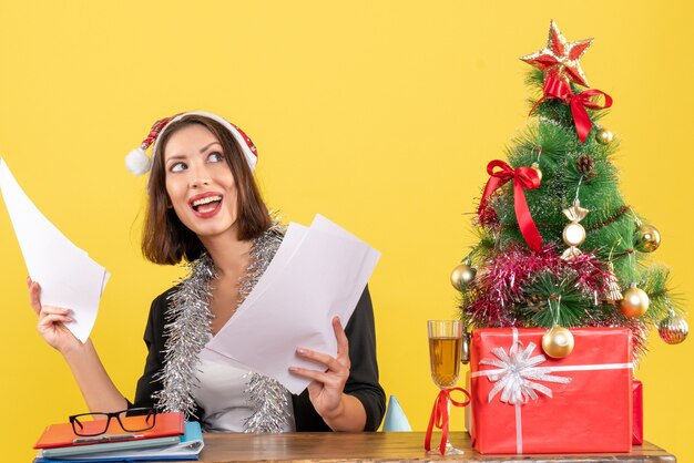 Mulher de negócios concentrada emocional satisfeita de terno com chapéu de Papai Noel e decorações de ano novo segurando documentos e sentada em uma mesa com uma árvore de natal no escritório