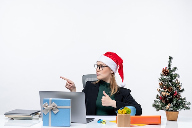 Mulher de negócios confiante com chapéu de papai noel sentada a uma mesa com uma árvore de natal e um presente nela fazendo um gesto de ok e apontando algo do lado direito no escritório sobre fundo branco