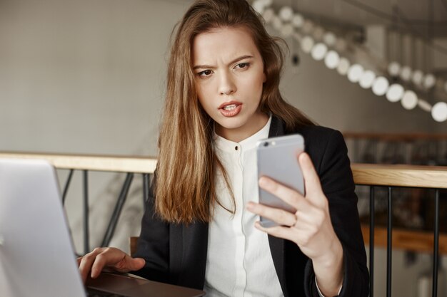 Mulher de negócios confusa e descontente olhando para o celular enquanto trabalha com o laptop no café