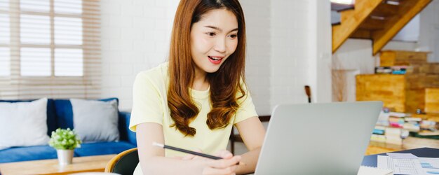 Mulher de negócios da Ásia usando laptop conversa com colegas sobre o plano de videochamada enquanto inteligente trabalhando em casa na sala de estar. Auto-isolamento, distanciamento social, quarentena para prevenção do vírus corona.